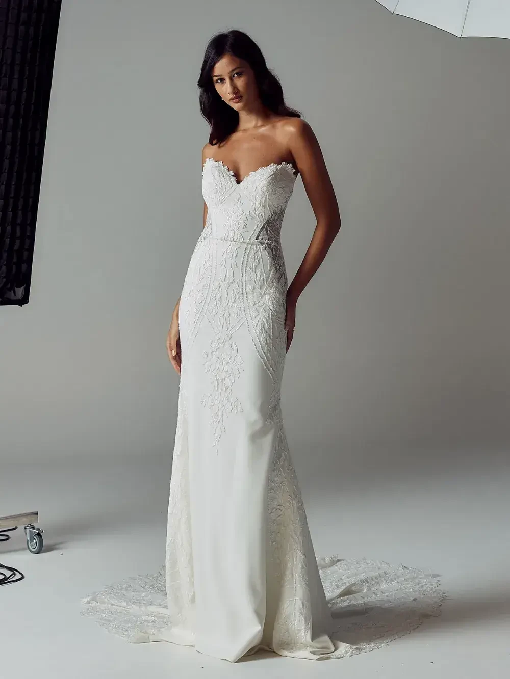 A woman wearing a strapless, white lace wedding gown with intricate floral patterns. She stands confidently in a studio setting, exuding elegance and grace.