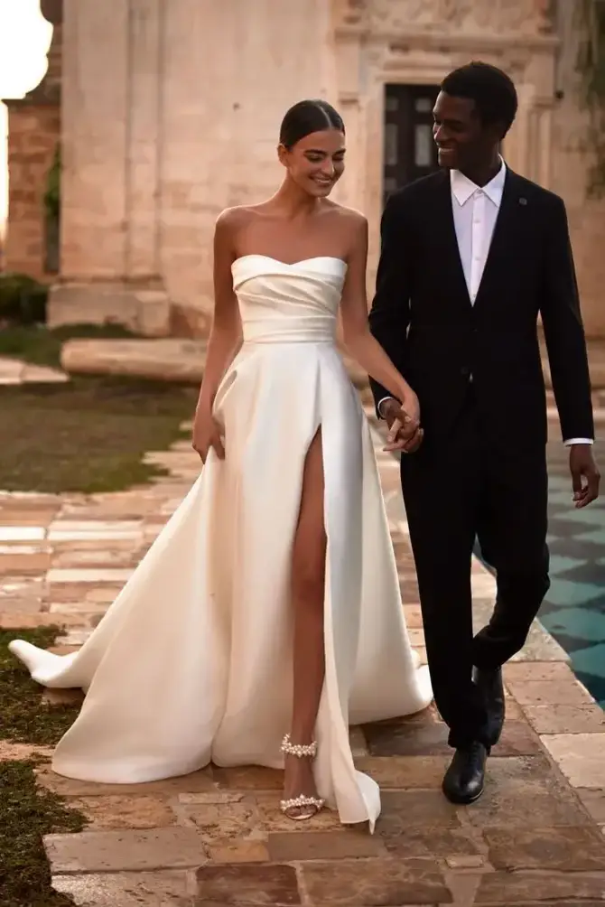 A joyful couple walks hand in hand outdoors. The woman wears an elegant, strapless white gown with a thigh-high slit, and the man is in a classic black suit.