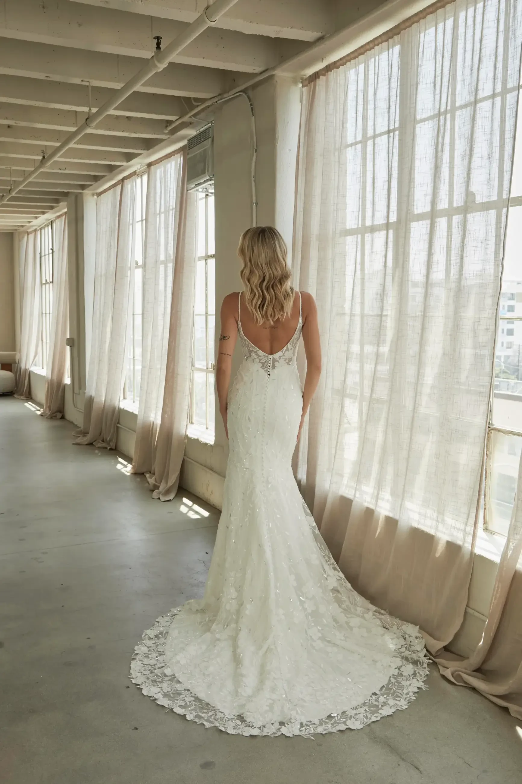 A woman in a lace wedding dress with a long train stands in a sunlit corridor with sheer curtains, creating an elegant and serene atmosphere.