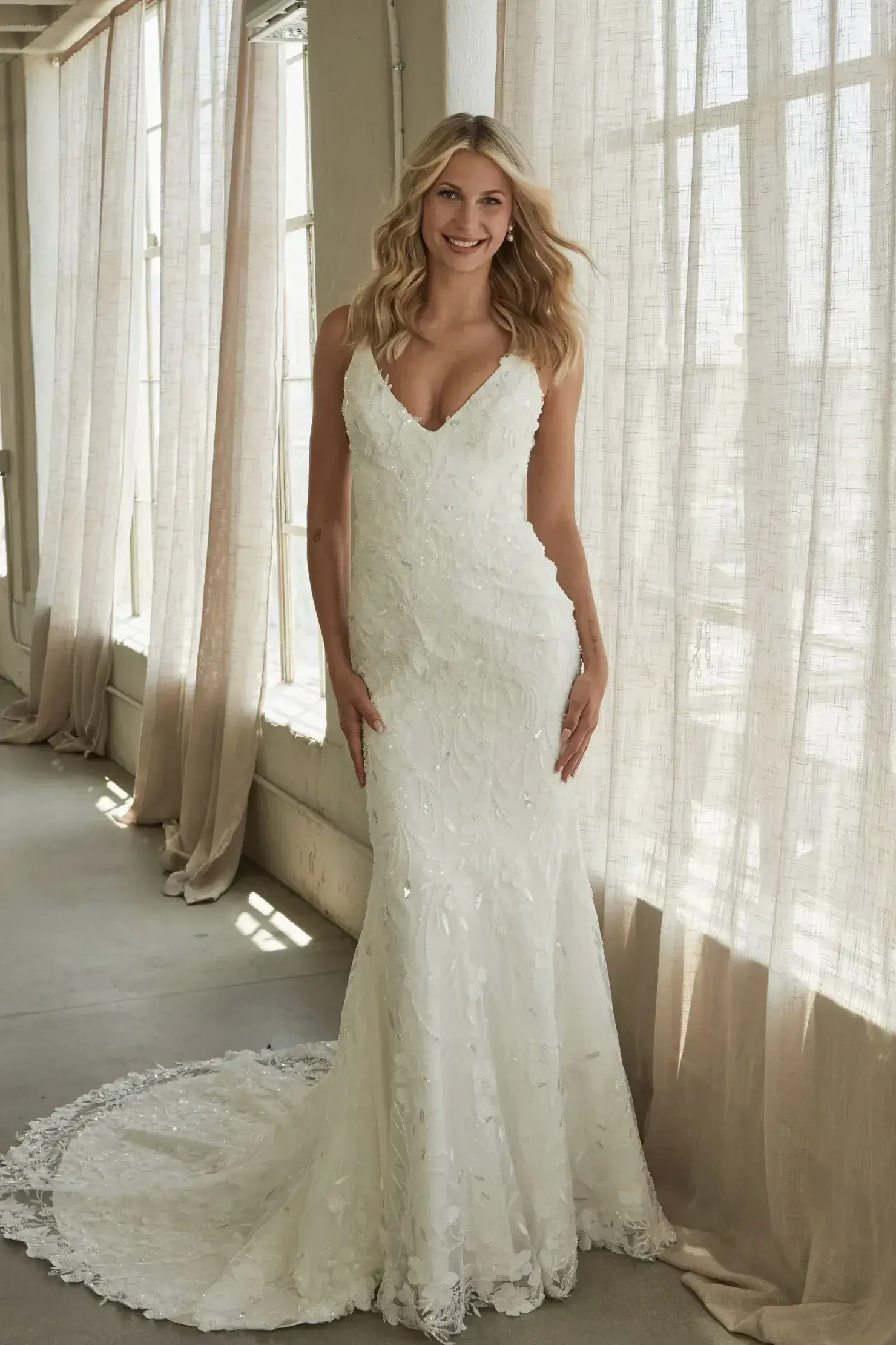 A woman smiles in an elegant white lace wedding gown with a long train, standing near large windows with sheer curtains, creating a soft, romantic ambiance.
