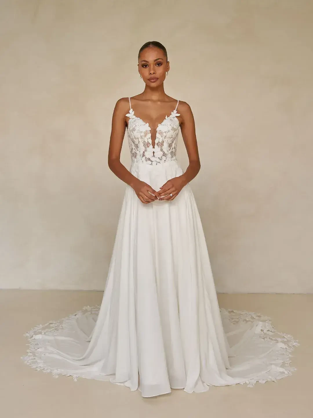 A woman stands poised in an elegant white gown with floral lace details and thin straps, exuding grace against a soft beige background.