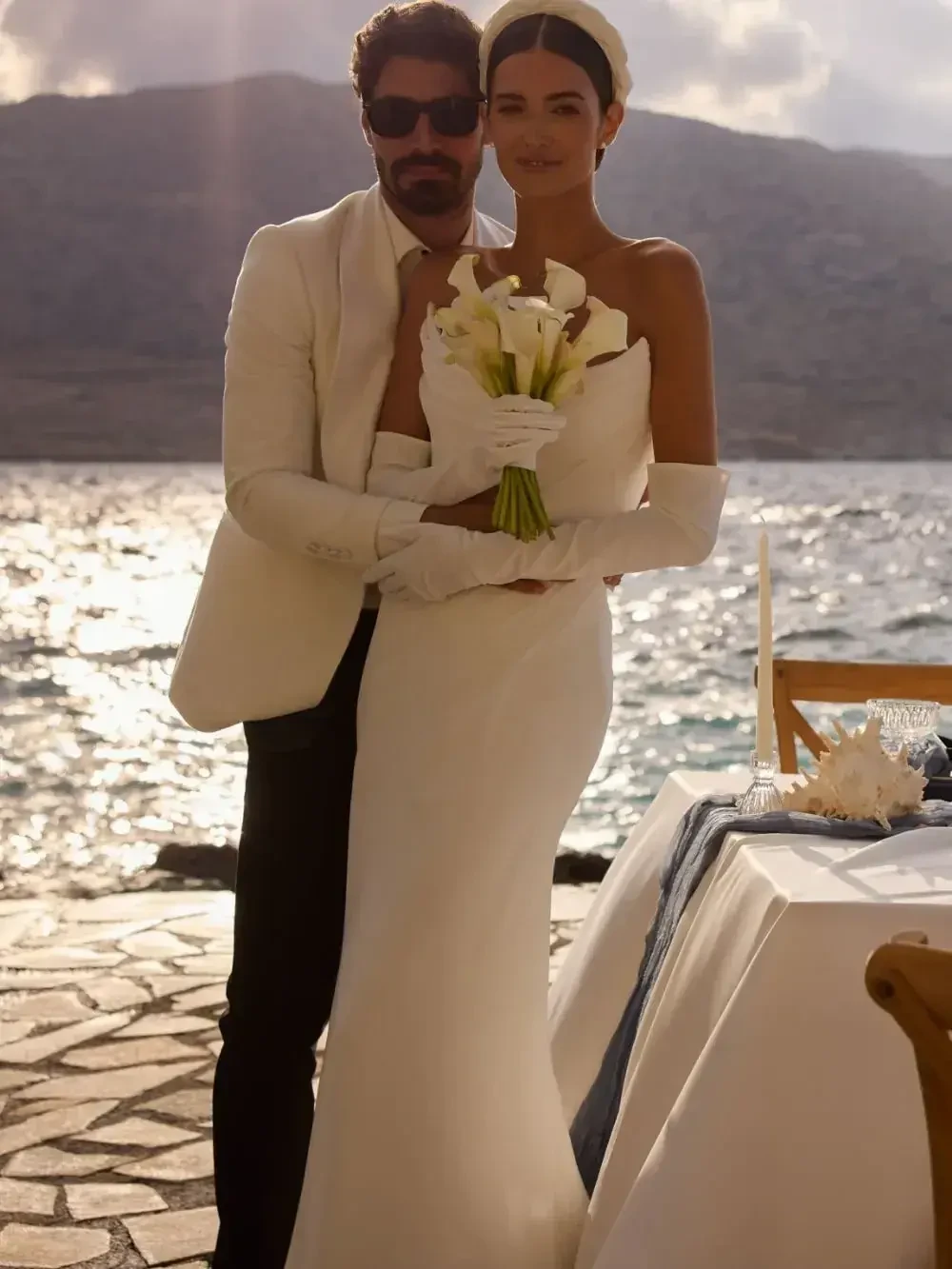 A bride in a white gown holding a bouquet of flowers stands beside a man dressed in a white suit, with a scenic waterfront background.