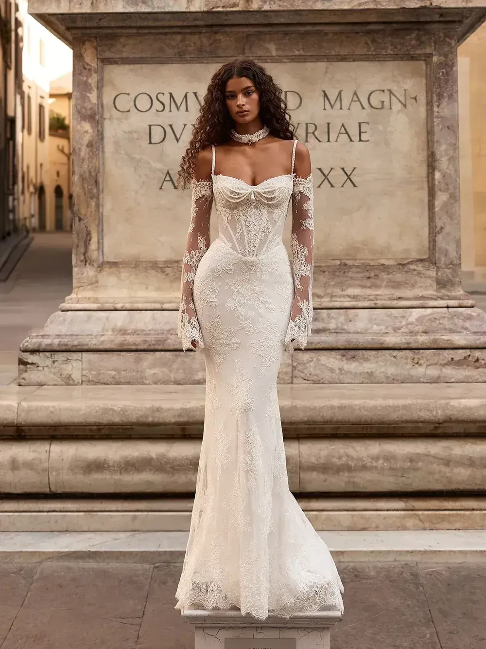 A model wearing an elegant white gown with intricate lace details, gloves, and a fitted silhouette, standing in an outdoor setting near a stone structure.
