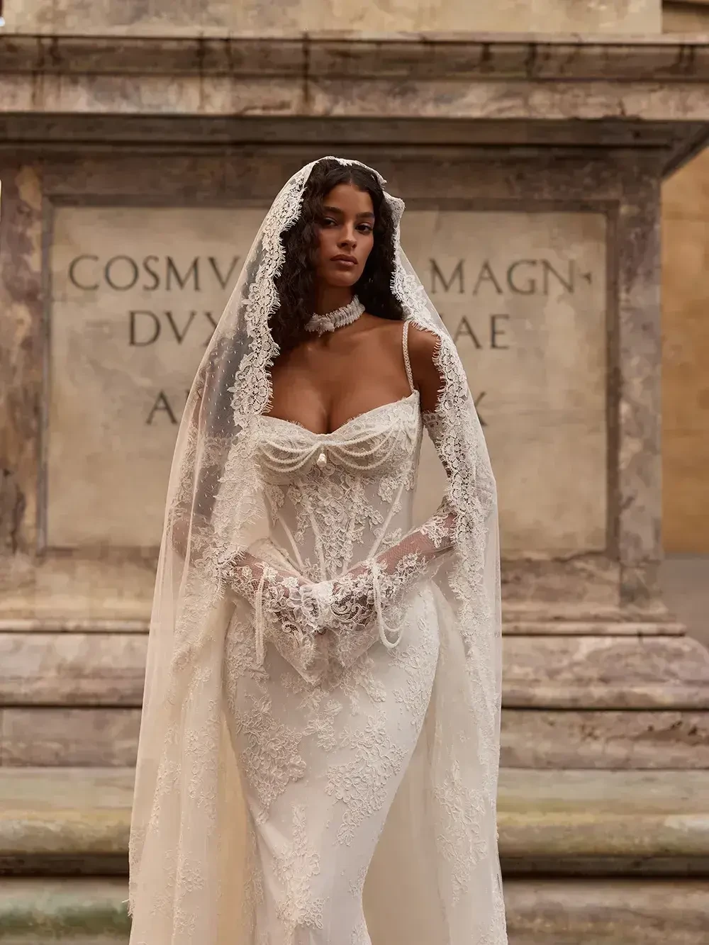 A model in an elegant wedding dress with intricate lace detailing and a long veil, standing in front of a stone backdrop.