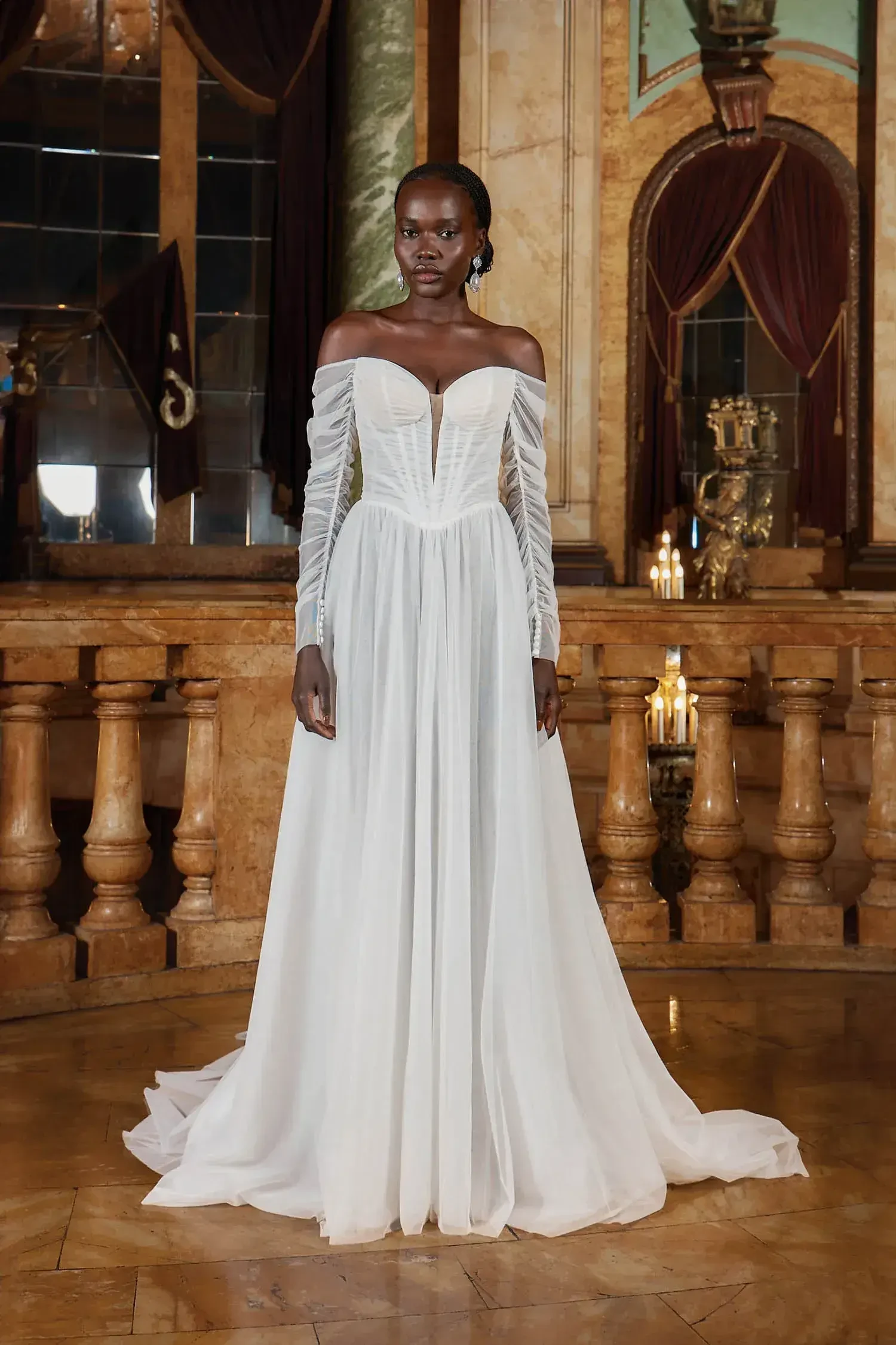 A model wearing a white off-the-shoulder gown with long sleeves, standing in an ornate indoor setting.