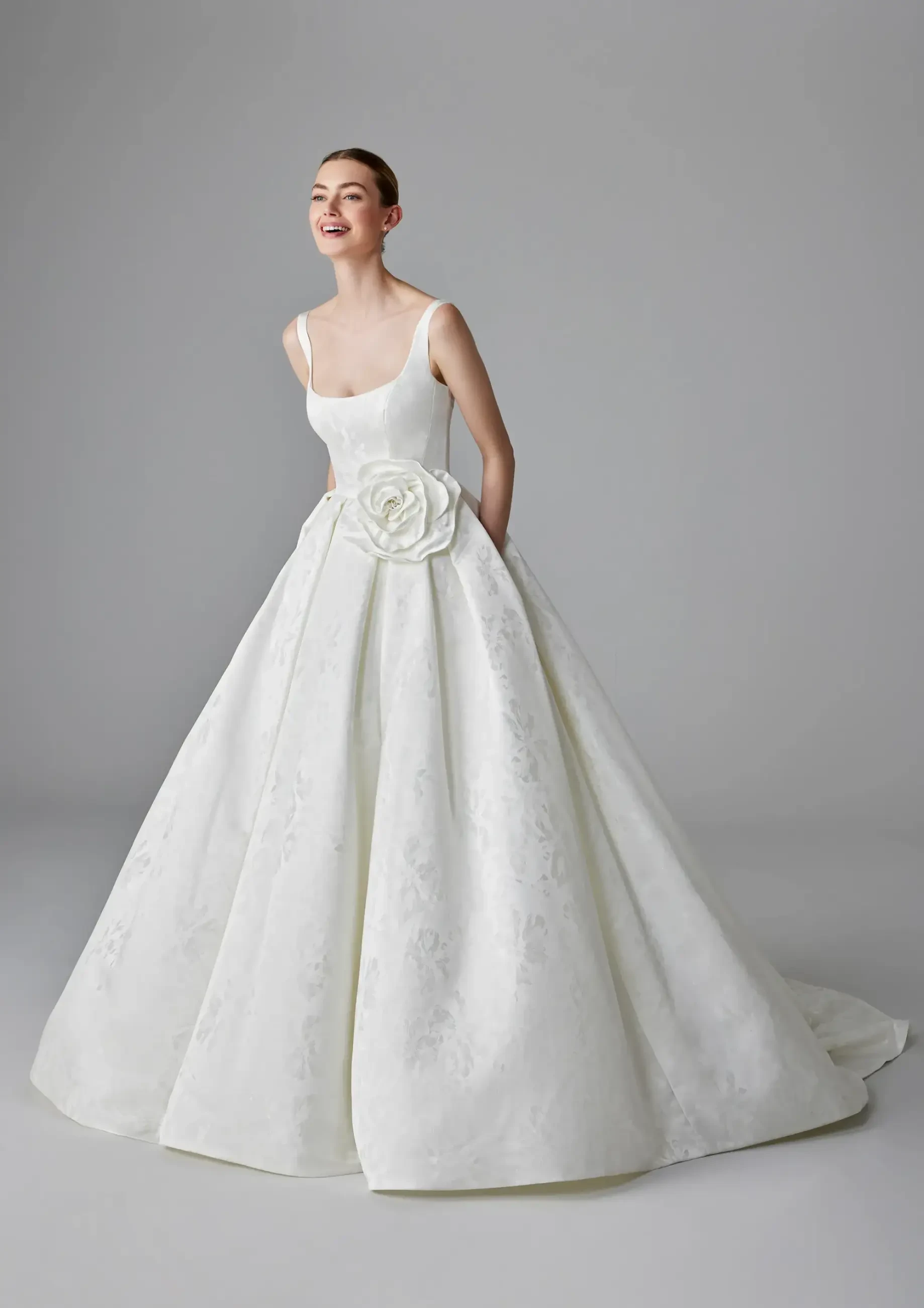 A joyful woman wearing an elegant white wedding gown with a floral embellishment at the waist, standing against a plain gray background.