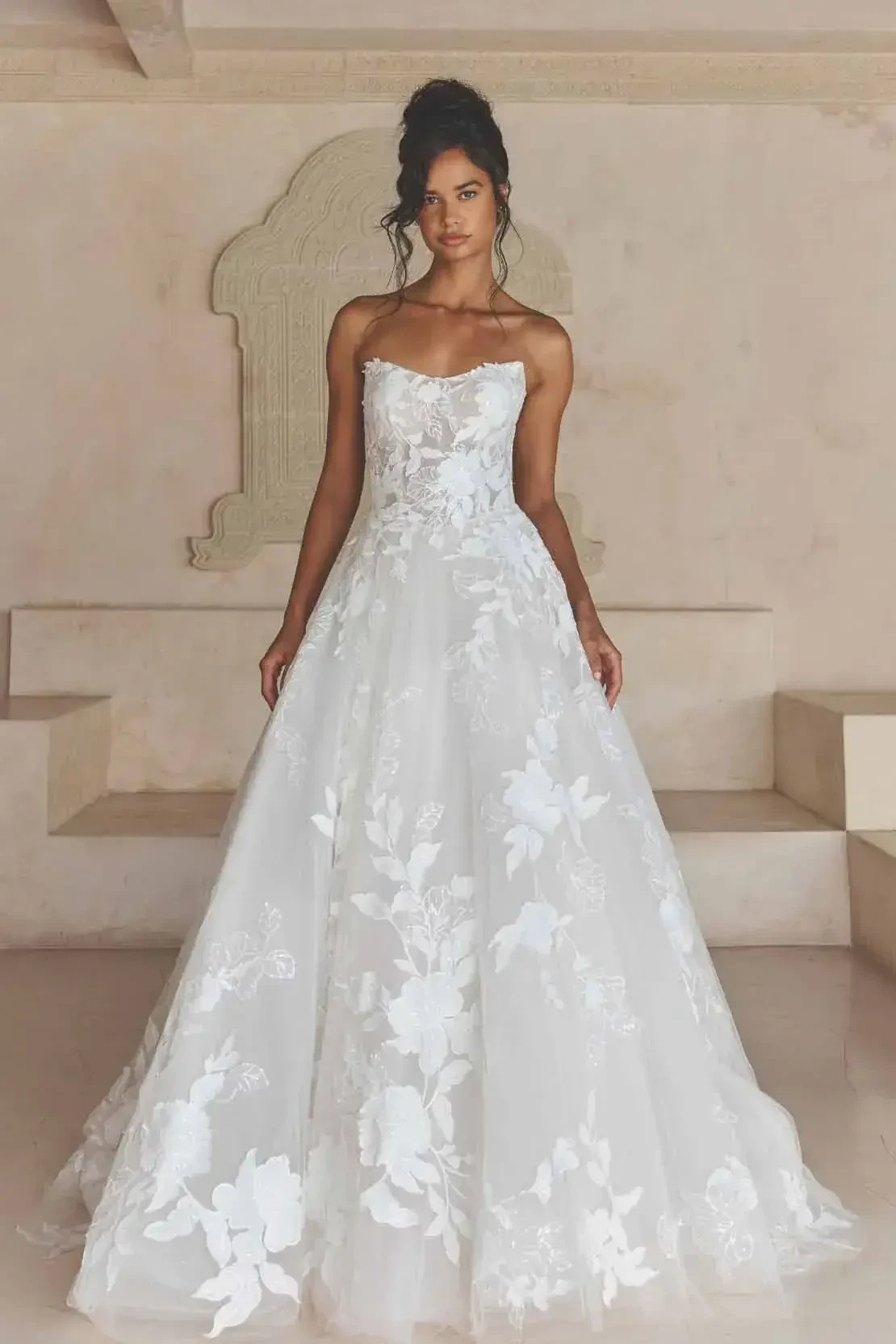 A model wearing a strapless wedding dress with floral embellishments, standing in a bright, minimalistic space.