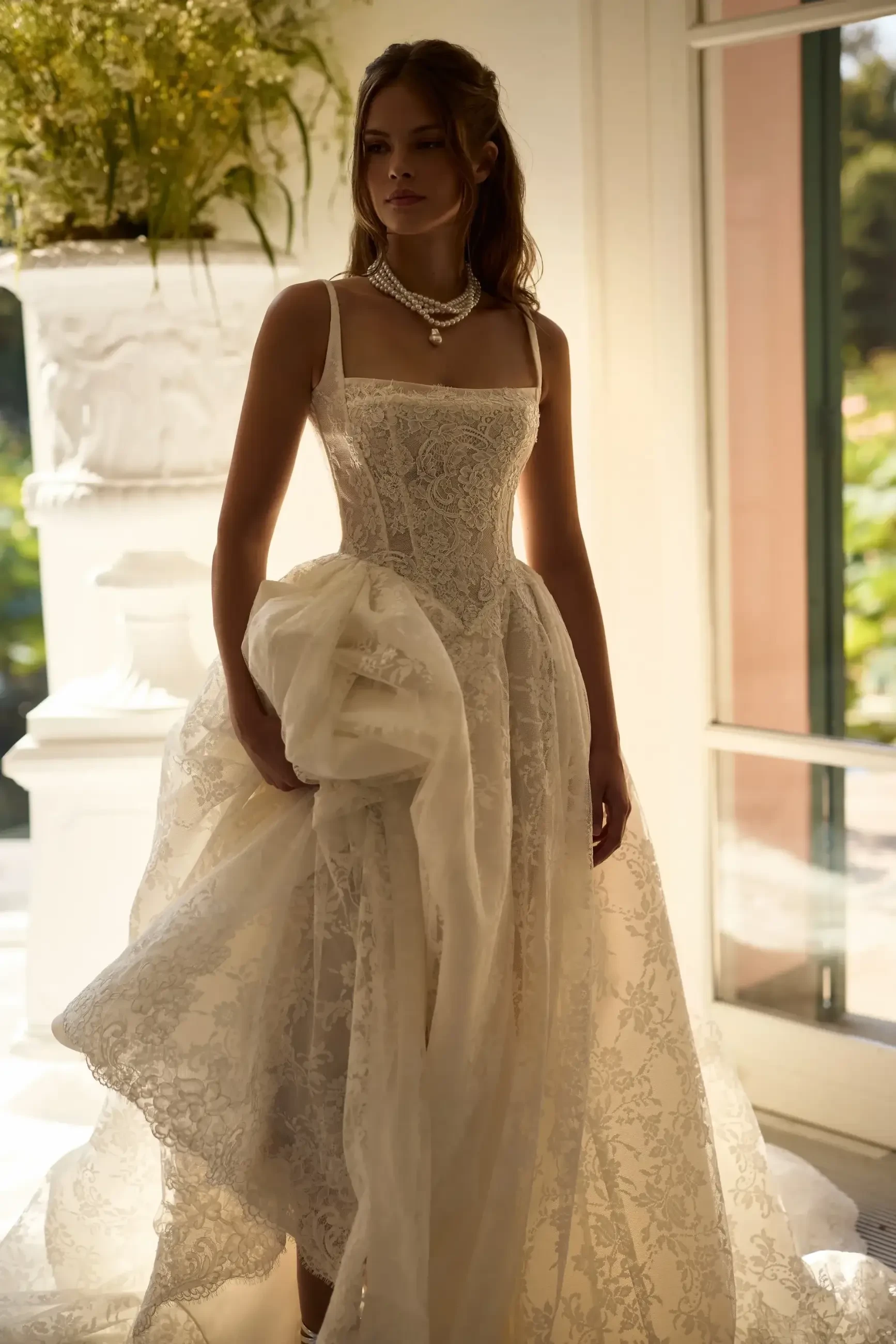 A woman in a beautiful intricately designed wedding dress, standing elegantly in a brightly lit room with greenery visible through the windows.
