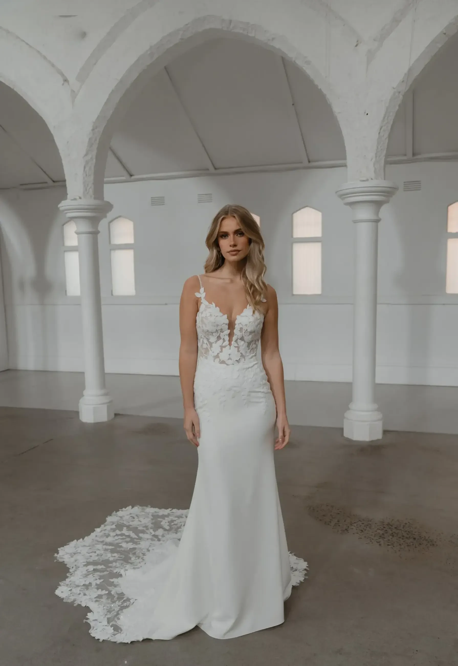 A woman wearing a white wedding dress stands in a spacious, architectural setting with arched columns and large windows.