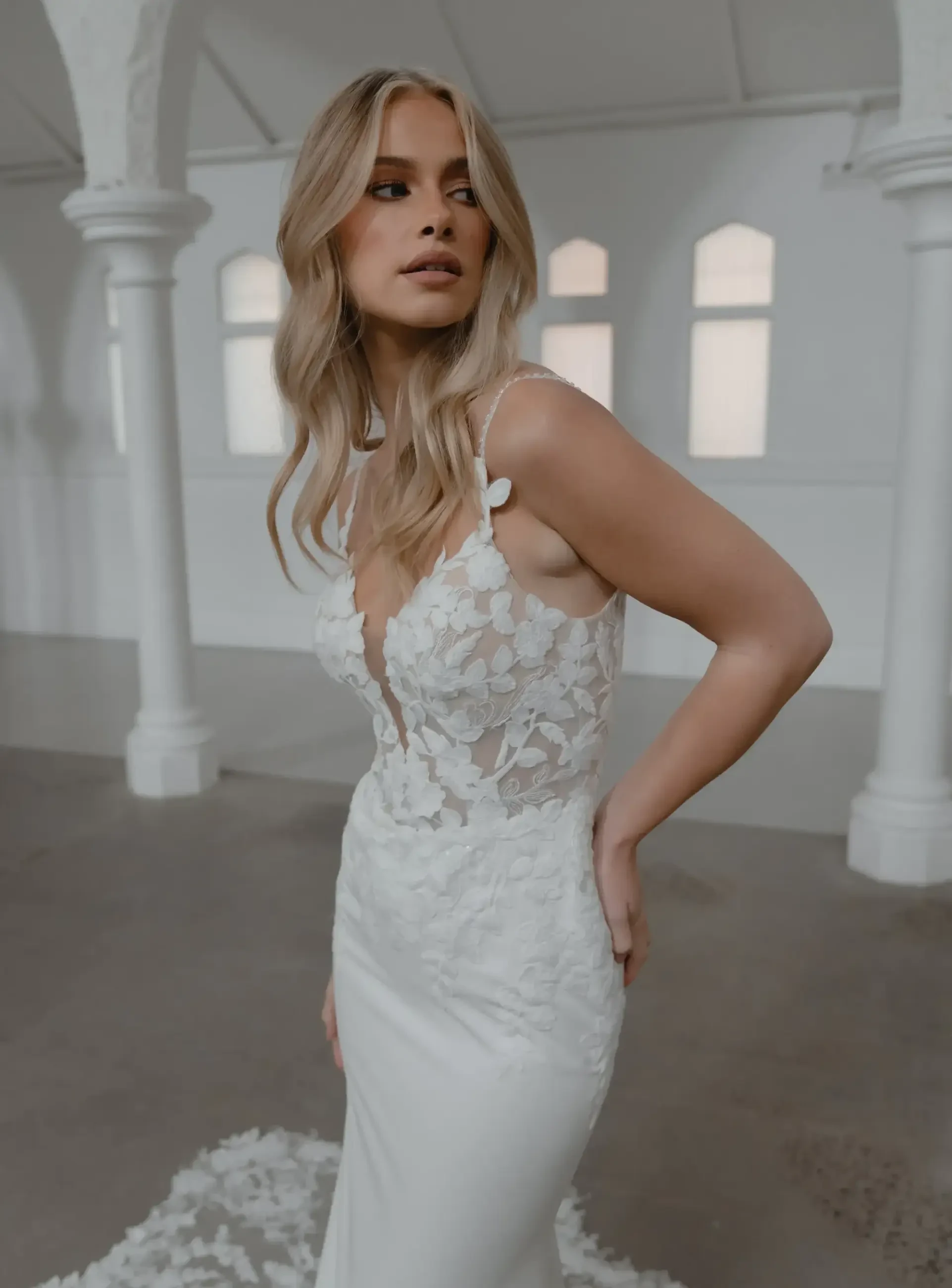 A woman in a white wedding dress with floral detailing, looking over her shoulder in an elegant venue.