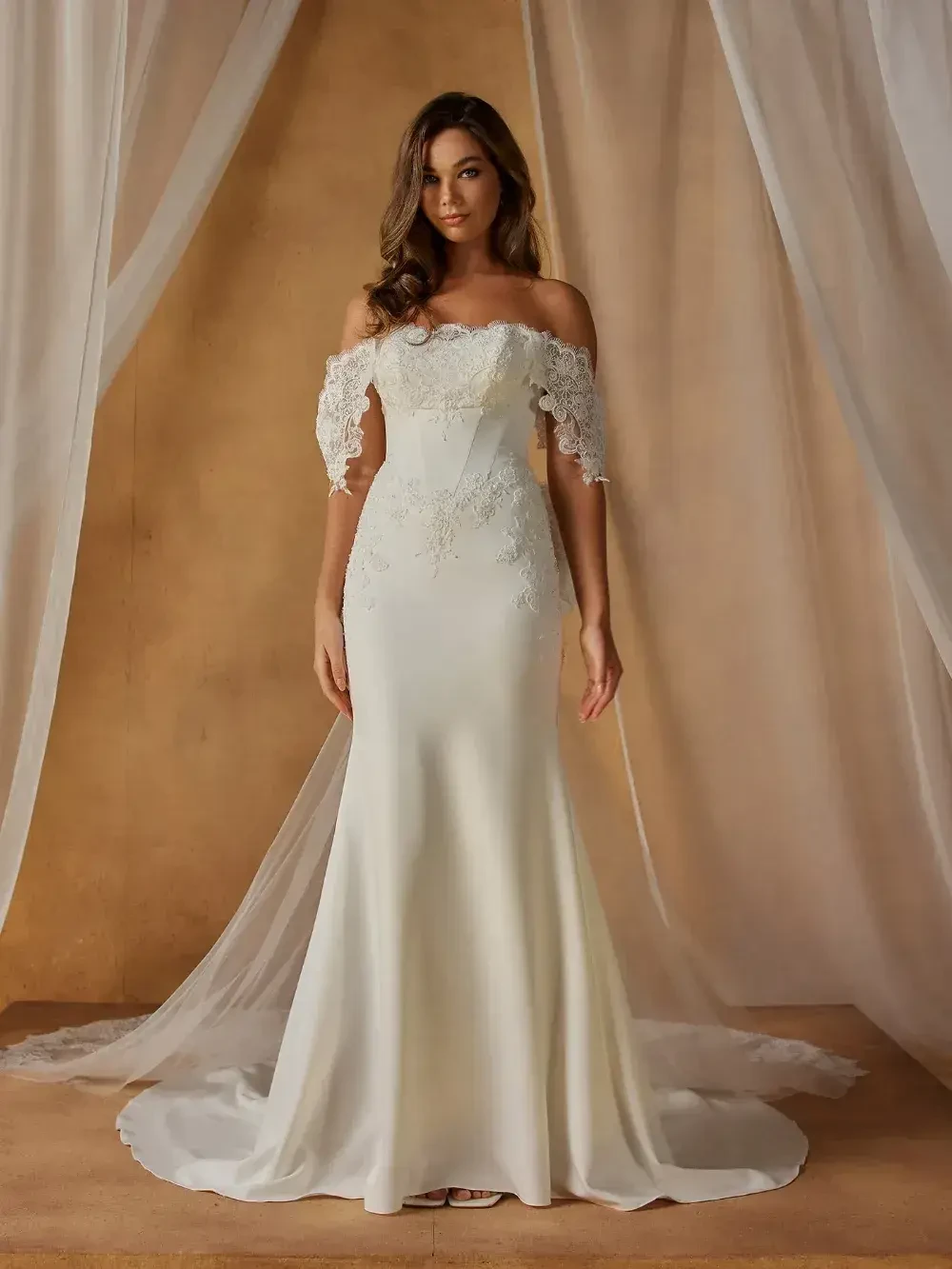 A woman wearing an elegant off-the-shoulder wedding dress with lace sleeves, standing in front of a backdrop of flowing fabric.