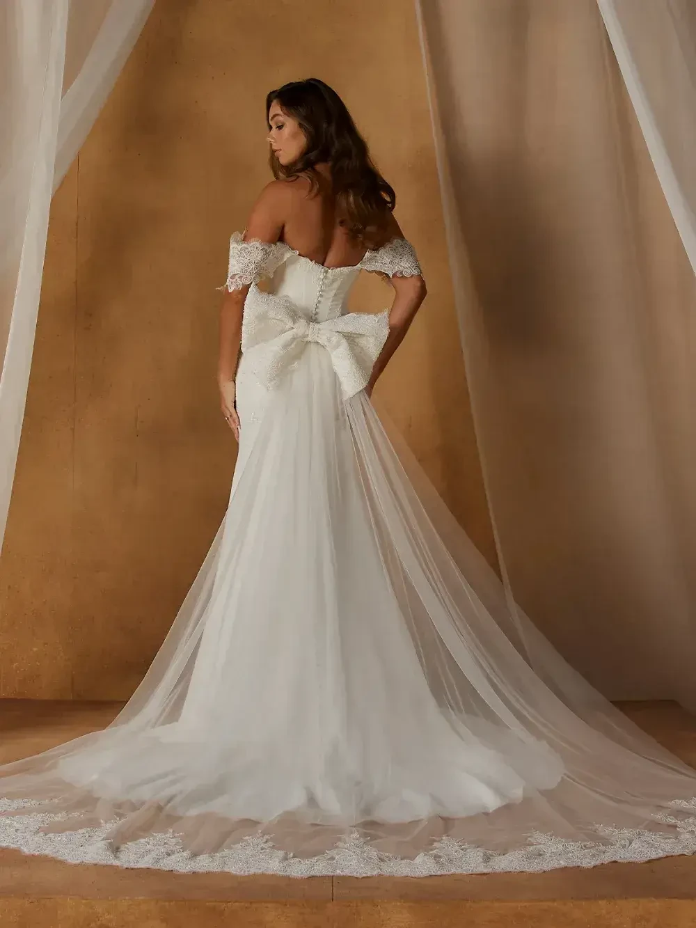 A woman is seen from behind wearing an elegant off-the-shoulder white wedding dress with a long train and a bow at the back. The background features soft, flowing fabric.