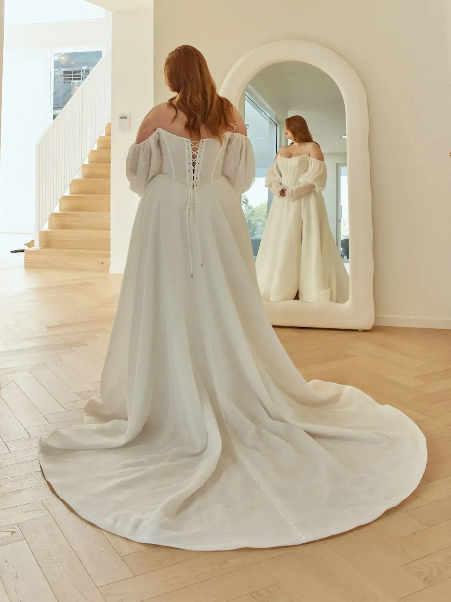 A person wearing a white gown with an off-the-shoulder design stands in front of a mirror, looking at their reflection. The gown has a flowing train and lace-up detail at the back. In the background, there is a staircase and bright natural light illuminating the space.
