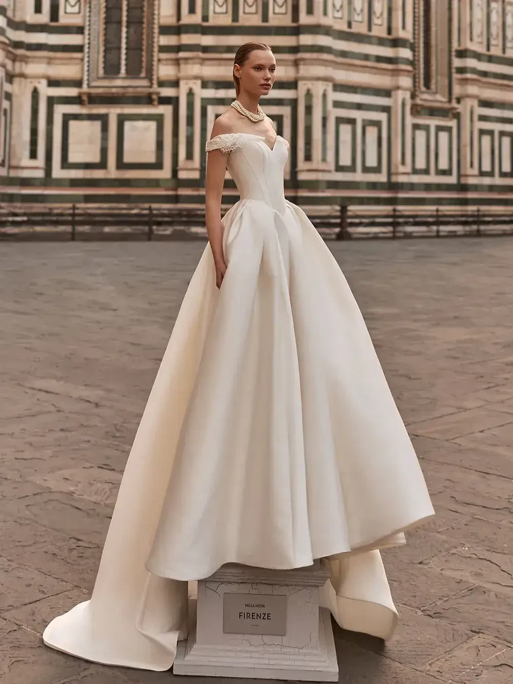 A model wearing a white wedding dress stands on a pedestal in a historic plaza with striking architecture in the background.
