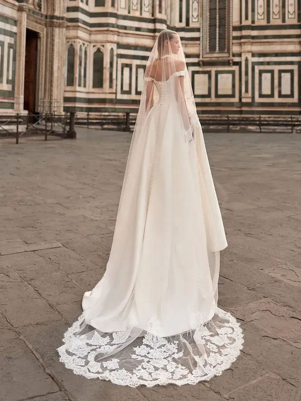 A woman in a flowing white wedding dress with a long veil stands in a cobblestone area, facing away from the camera, with a historic building in the background.