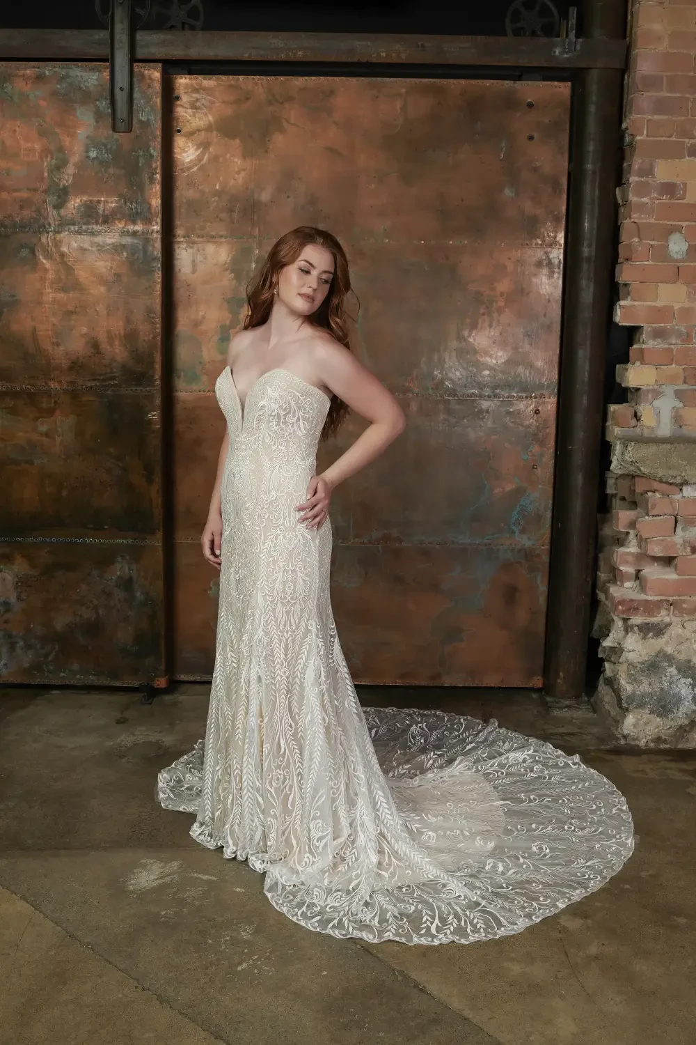 A woman poses in a shimmering, fitted wedding dress with a long train, standing against a rustic, copper-colored backdrop.